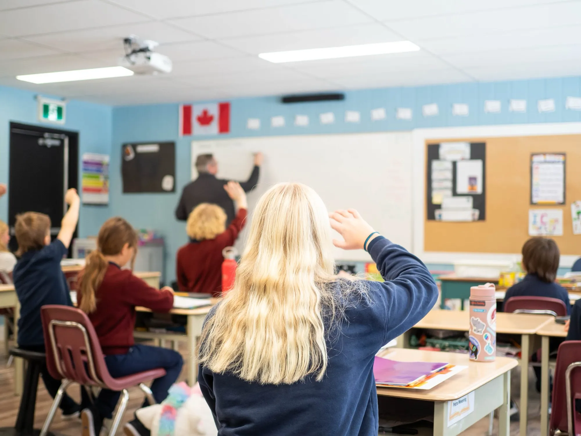 Elementary students raising their hands while a teacher writes on the whiteboard