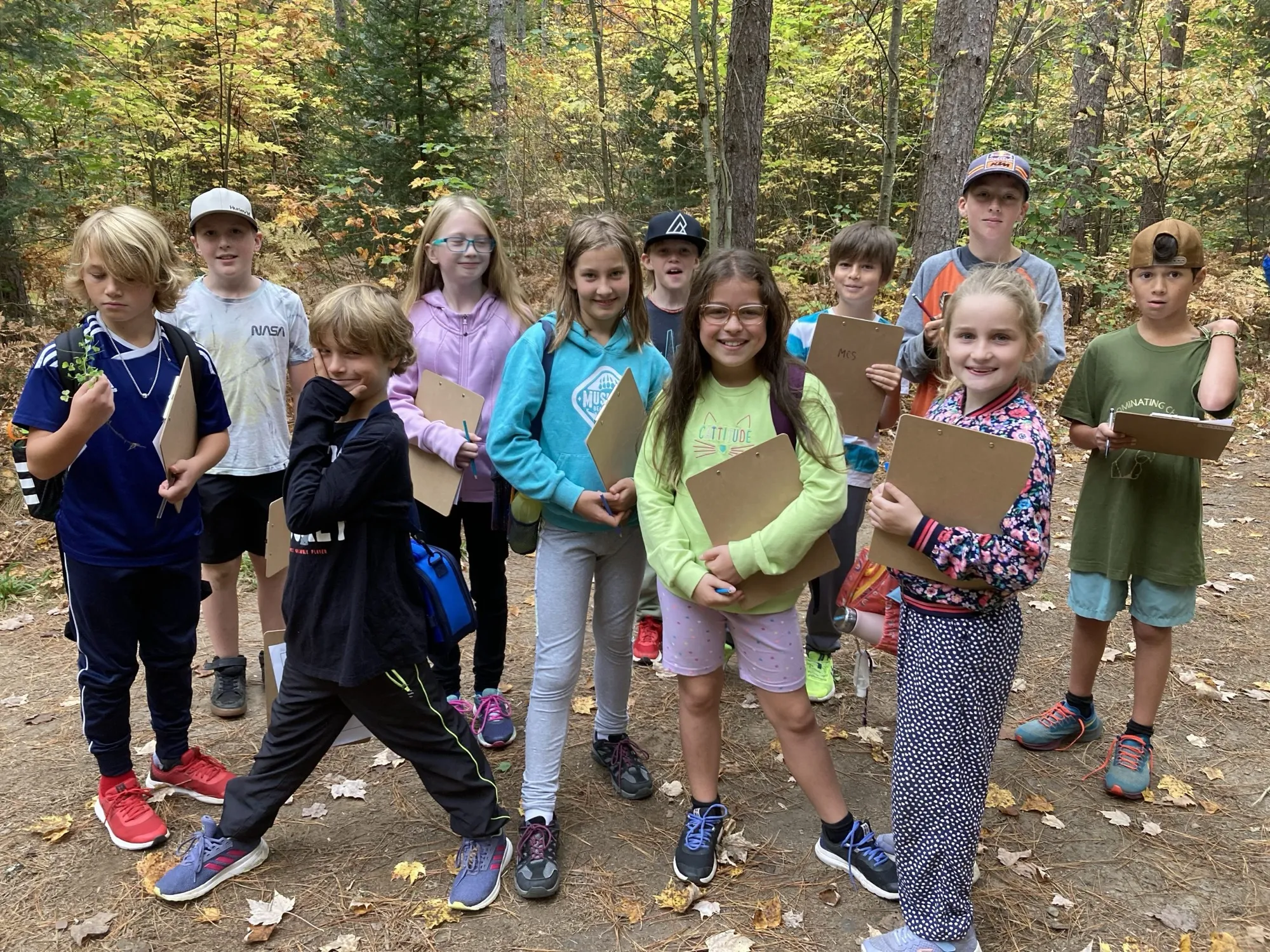 A class of elementary students with clipboards gathered in a fall forest during a Forest School day