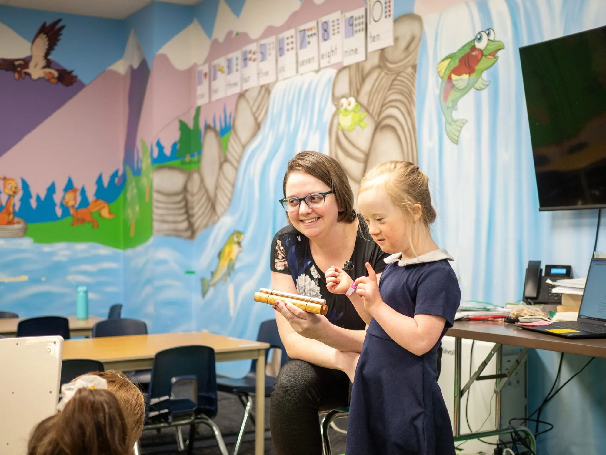 A smiling teacher and a young student sharing a classroom activity in front of a painted mural