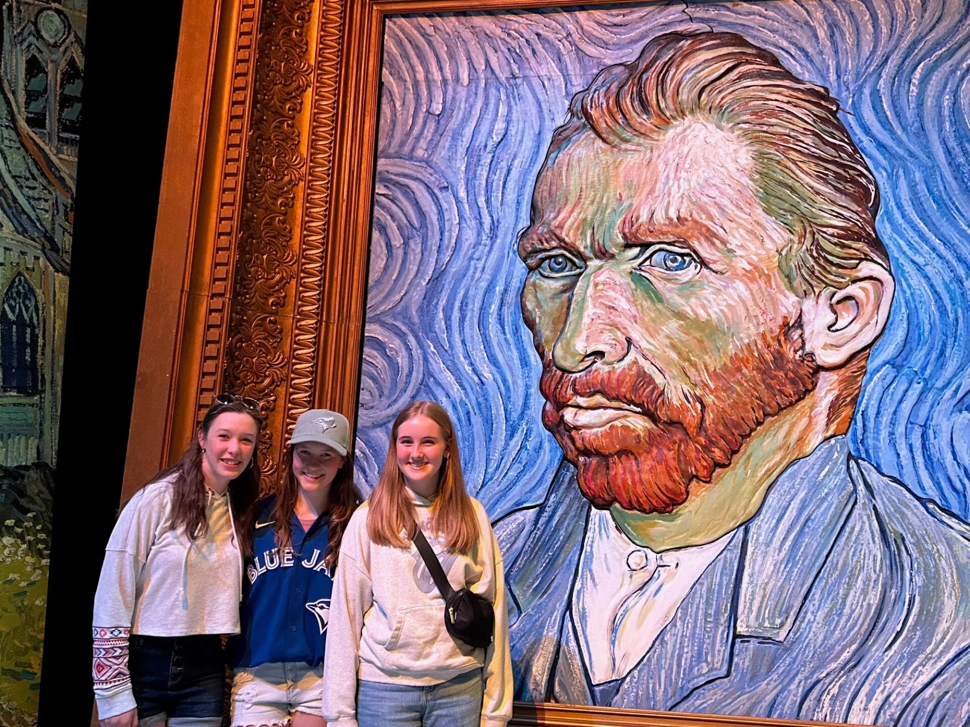 Three students posing beside a large Van Gogh portrait at an art exhibit